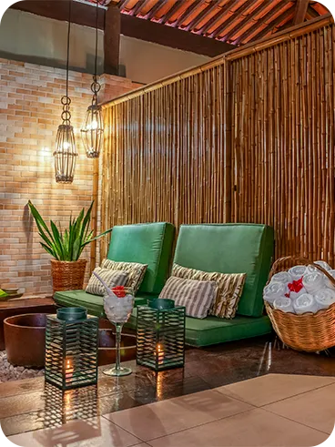 Espaço de spa com decoração aconchegante e rústica, poltronas verdes, cestas com toalhas enroladas, iluminação suave com velas e luminárias pendentes, parede de bambu e plantas decorativas