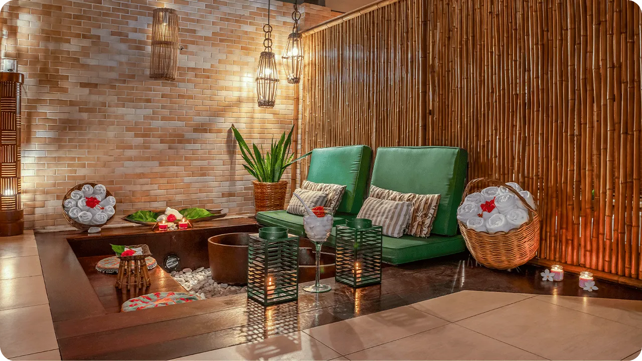 Espaço de spa com decoração aconchegante e rústica, poltronas verdes, cestas com toalhas enroladas, iluminação suave com velas e luminárias pendentes, parede de bambu e plantas decorativas
