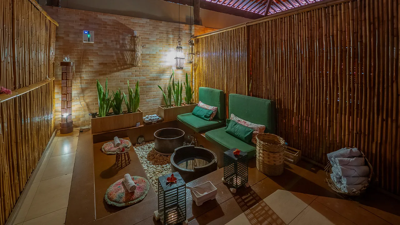 Espaço de SPA com decoração aconchegante e rústica, poofs verdes em cima do banco de madeira, cestas com toalhas enroladas, iluminação suave com velas e luminárias pendentes, parede de bambu e plantas decorativas.