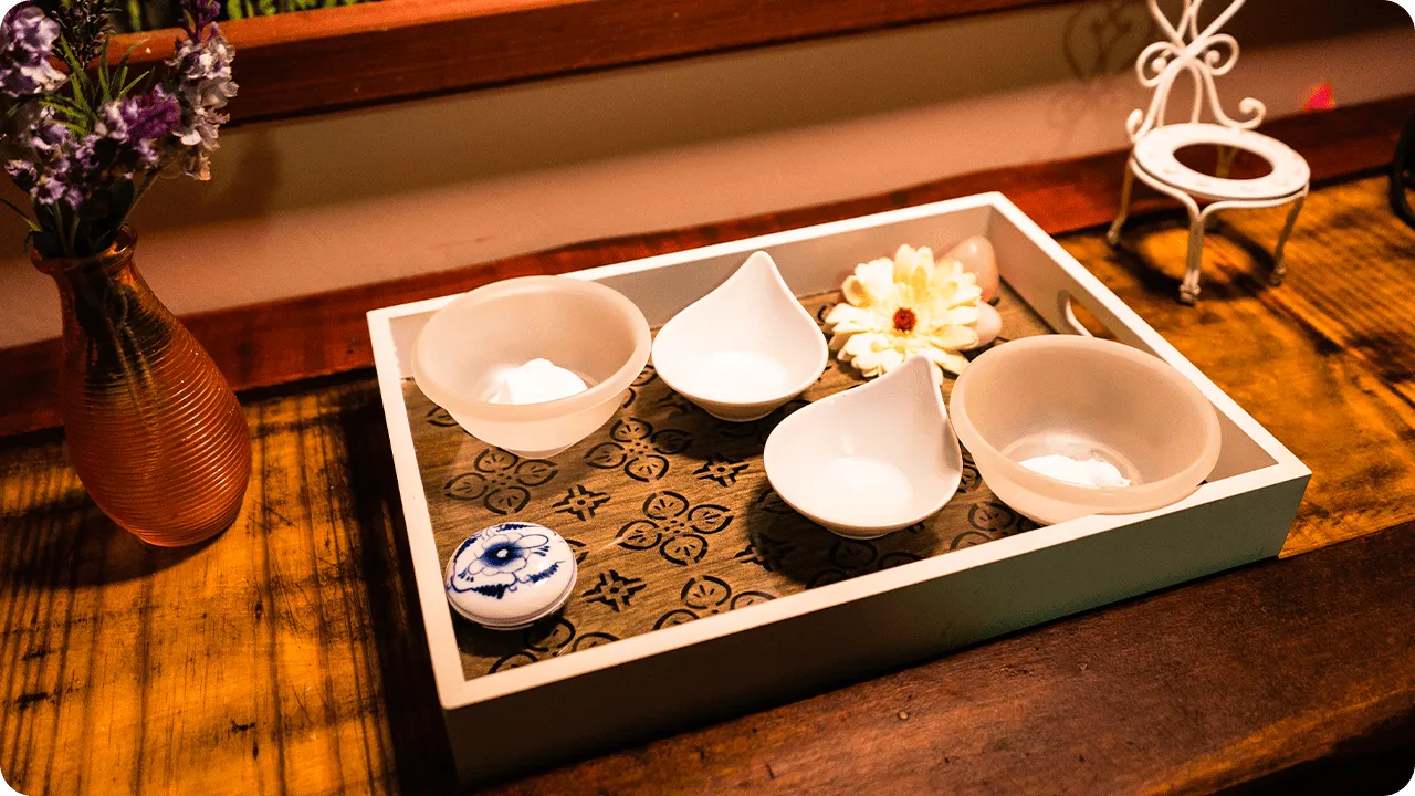 Decorative tray on a rustic wooden table with white ceramic pots, artificial flower, decorative soap, mini white iron chair and amber vase with lilac flowers.