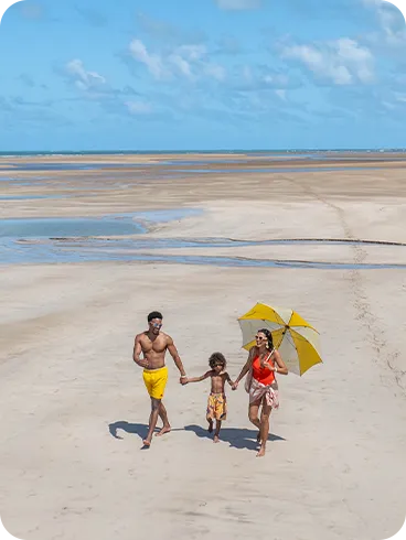 Família caminhando em praia de maré baixa, com areia clara, céu azul e vegetação de coqueiros ao fundo