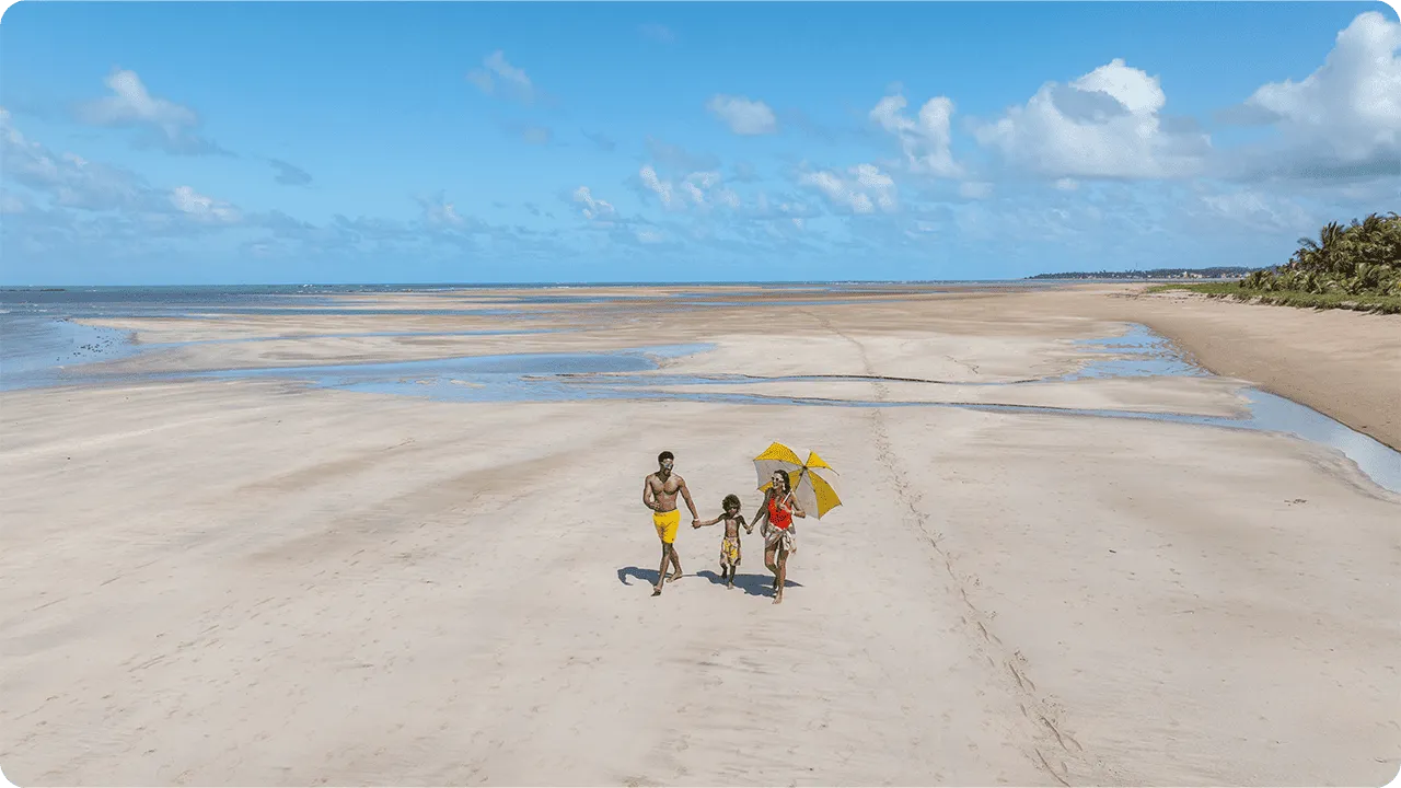Família caminhando em praia de maré baixa, com areia clara, céu azul e vegetação de coqueiros ao fundo