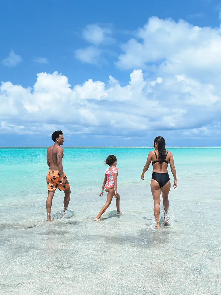 Família caminhando no extenso banco de areia do Caminho de Moisés, em Maragogi, durante a maré baixa. A paisagem mostra o mar calmo e azul-turquesa de ambos os lados da faixa de areia, sob um céu azul amplo.