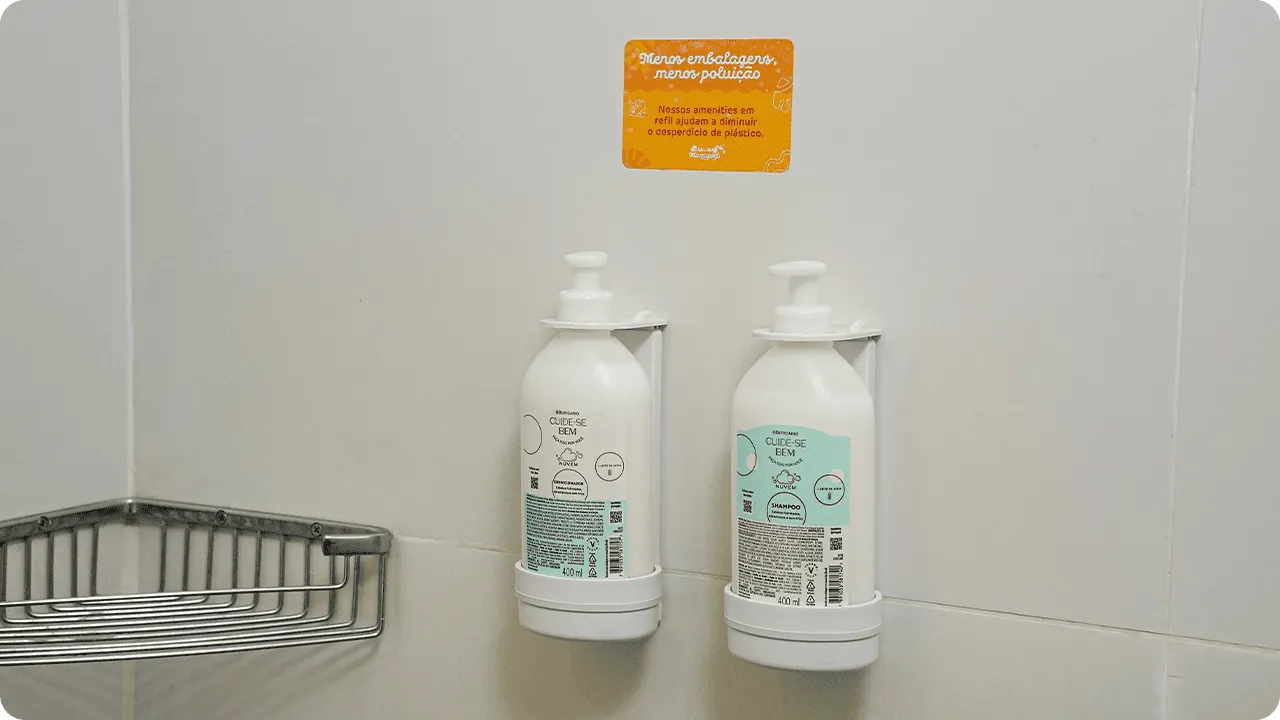 Shampoo e condicionador O Boticário em dispensers de refil na parede do banheiro, acompanhados de um aviso sobre sustentabilidade e redução de plástico.