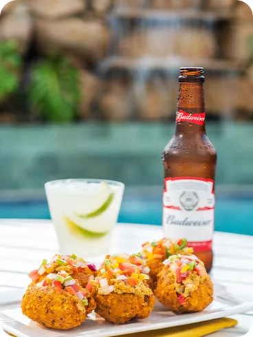 Prato com bolinhos crocantes cobertos por vinagrete ao lado de uma cerveja Budweiser e um drink cítrico com gelo sobre mesa branca; ao fundo, cascata e piscina do Salinas Maragogi.