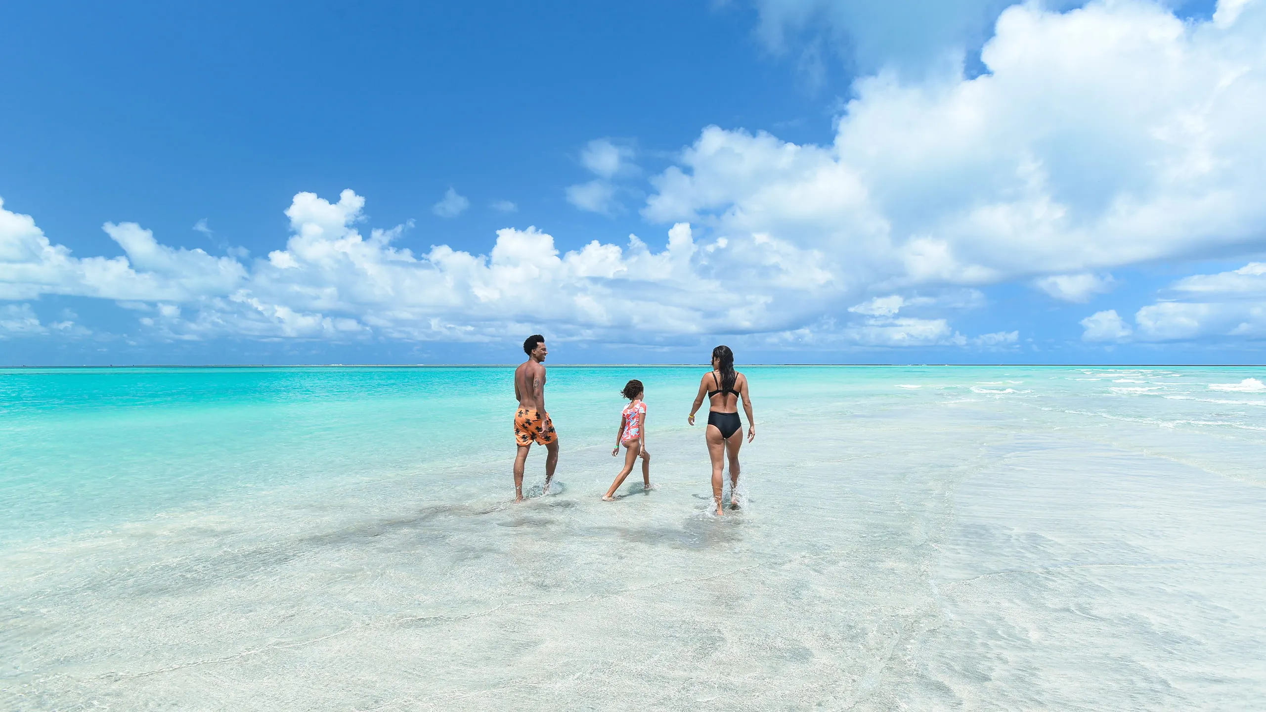 Família caminhando no extenso banco de areia do Caminho de Moisés, em Maragogi, durante a maré baixa. A paisagem mostra o mar calmo e azul-turquesa de ambos os lados da faixa de areia, sob um céu azul amplo.