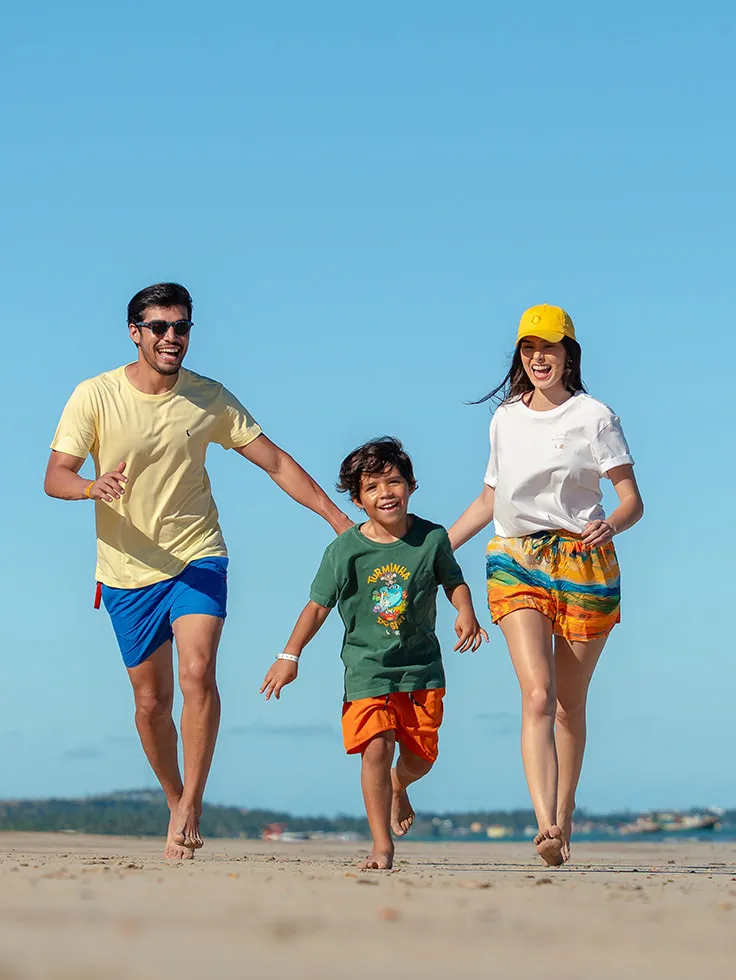 Família correndo na areia da praia em dia ensolarado e sem nuvens. O pai está ao lado esquerdo, o filho no meio e a mãe do lado direito da imagem.