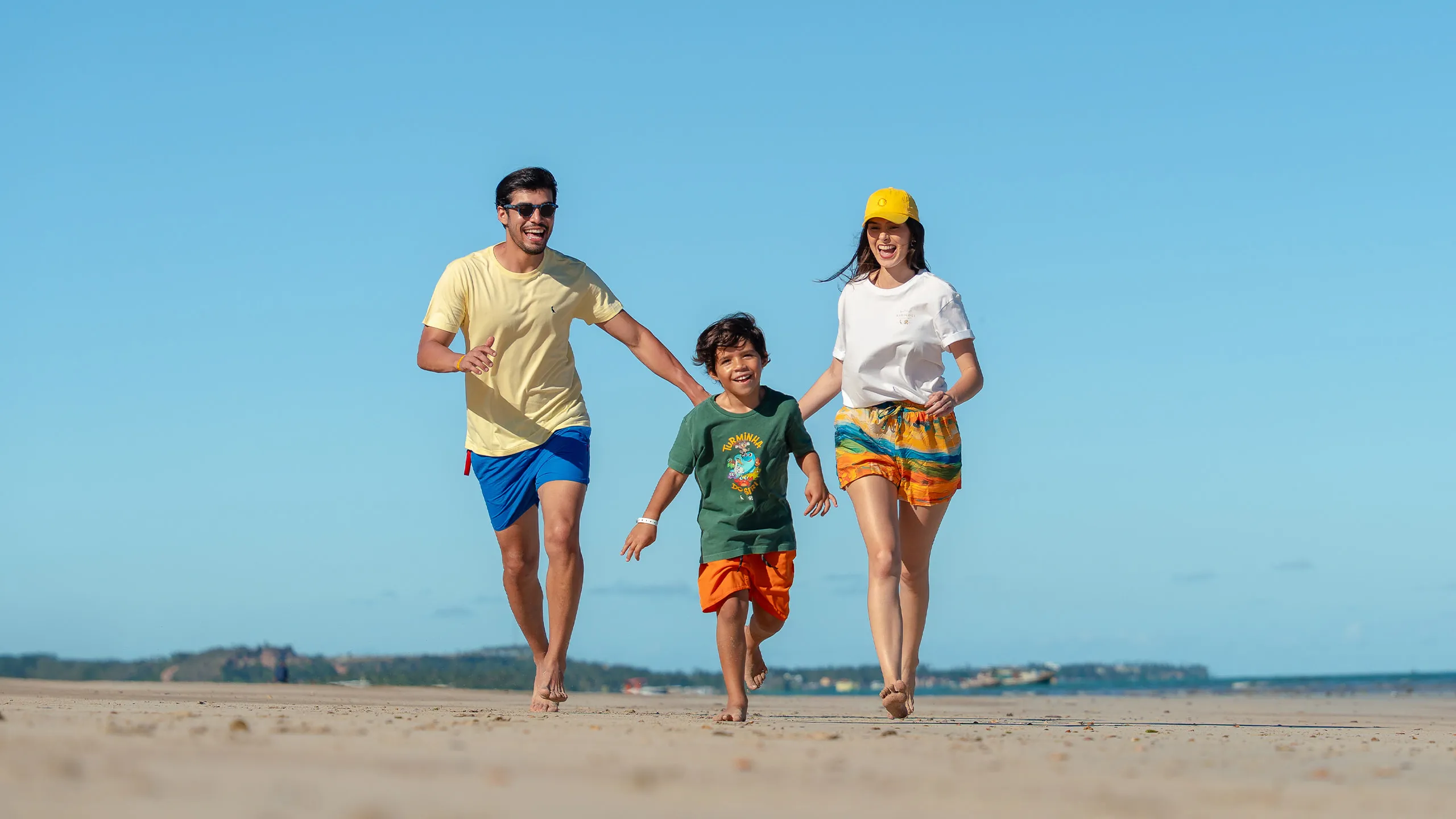 Família correndo na areia da praia em dia ensolarado e sem nuvens. O pai está ao lado esquerdo, o filho no meio e a mãe do lado direito da imagem.