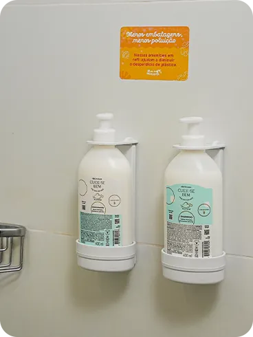 Shampoo e condicionador O Boticário em dispensers de refil na parede do banheiro, acompanhados de um aviso sobre sustentabilidade e redução de plástico.