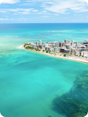Vista aérea de la Playa Ponta Verde en Maceió, que destaca el mar cristalino con múltiples tonos de azul y verde, y la costa bordeada de edificios y hoteles bajo un cielo azul.