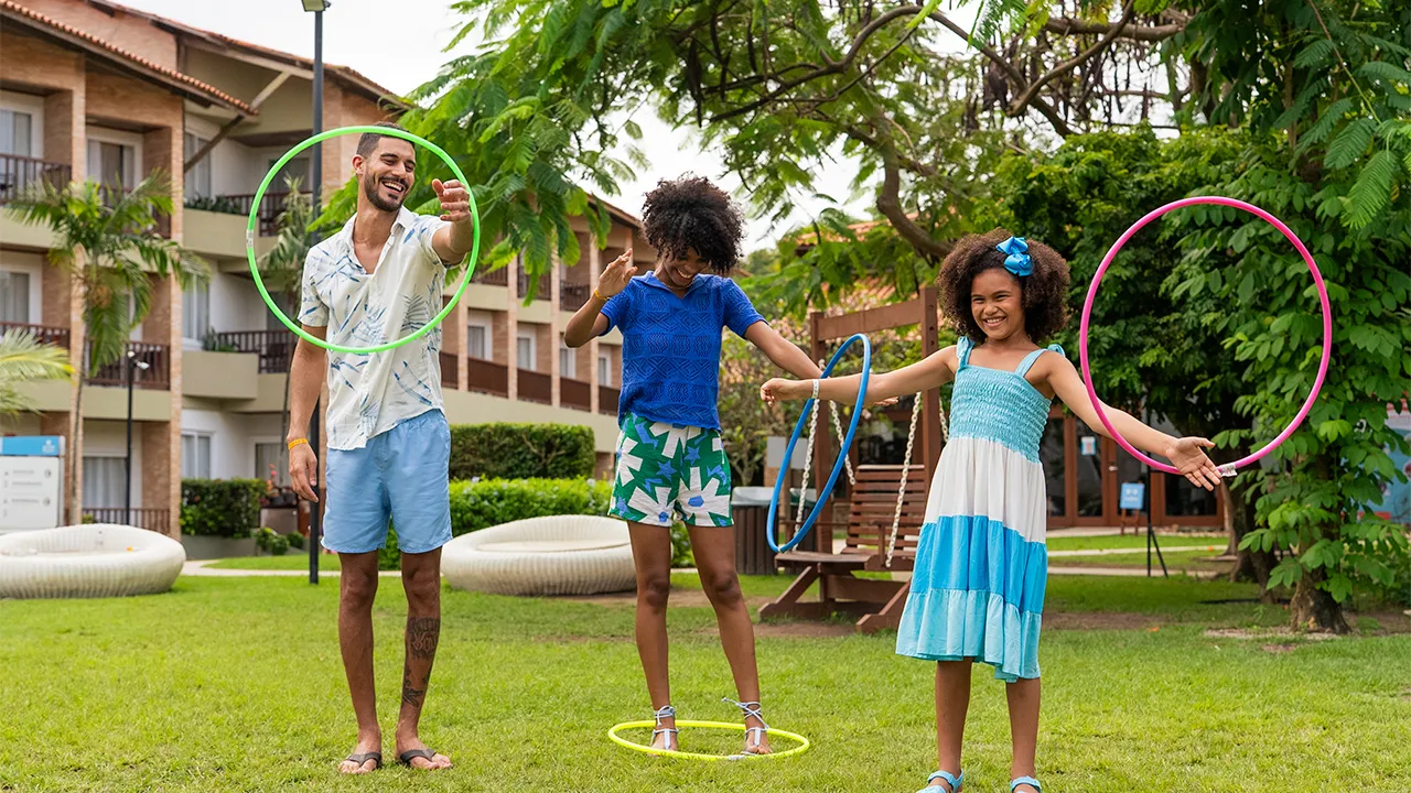 Família brincando com bambolê na área de lazer no Salinas Maceió. A filha está com dois bambolê e os pais cada um com um bambolê, no fundo tem a estrutura do resort e eles estão num gramado.