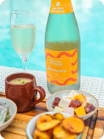Una botella de vino blanco espumoso Moscatel Salinas Maragogi junto a una copa y aperitivos, incluyendo una porción de camarones, queso en cubos y ceviche, con la piscina de fondo.