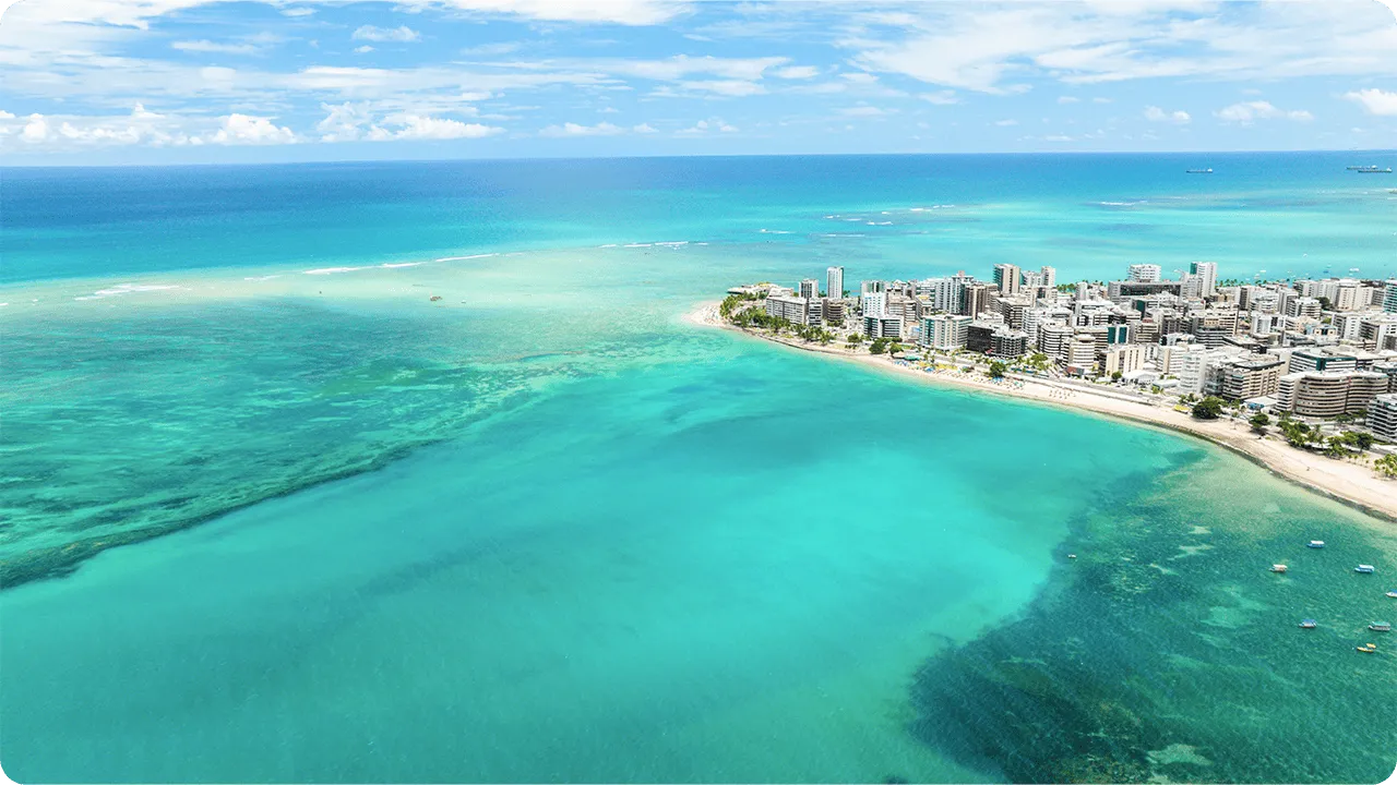 Vista aérea de la Playa Ponta Verde en Maceió, que destaca el mar cristalino con múltiples tonos de azul y verde, y la costa bordeada de edificios y hoteles bajo un cielo azul.