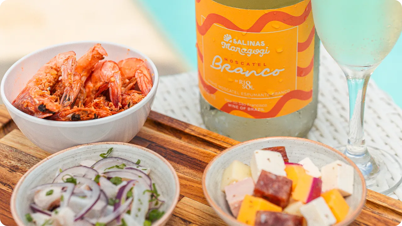 Una botella de vino blanco espumoso Moscatel Salinas Maragogi junto a una copa y aperitivos, incluyendo una porción de camarones, queso en cubos y ceviche, con la piscina de fondo.