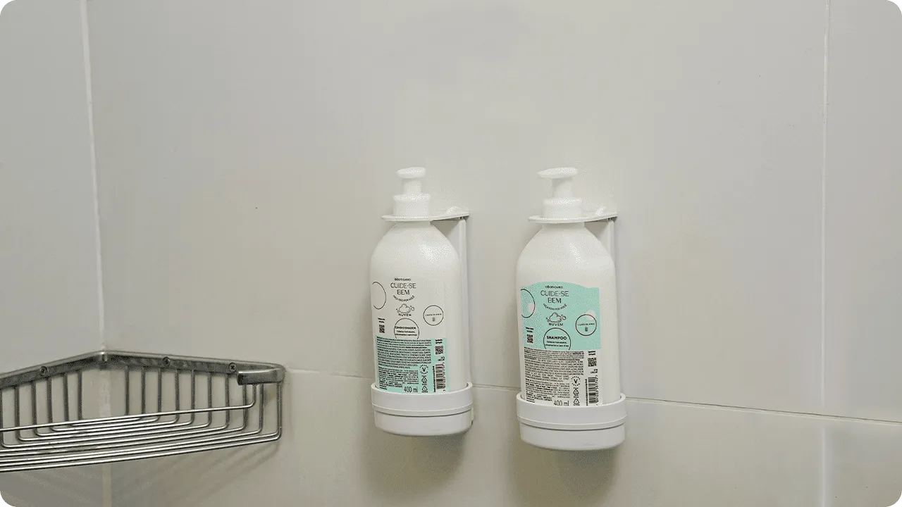 Shampoo e condicionador O Boticário, da linha Cuide-se Bem, em frascos de refil instalados em dispensers na parede de azulejos do banheiro.