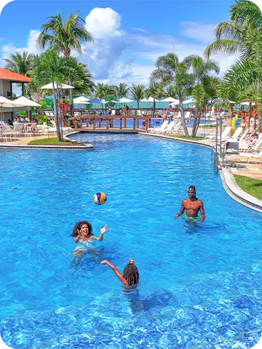 Una familia sonriente juega voleibol en la piscina azul de Salinas Maceió. La madre lanza la pelota al aire mientras padre e hija observan, en un día soleado rodeados de otros huéspedes y cocoteros.