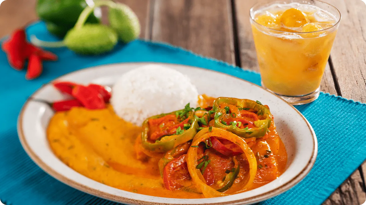 Prato de moqueca com arroz branco, molho cremoso alaranjado e rodelas de pimentão e tomate, acompanhado de caipirinha em copo baixo sobre toalha azul.