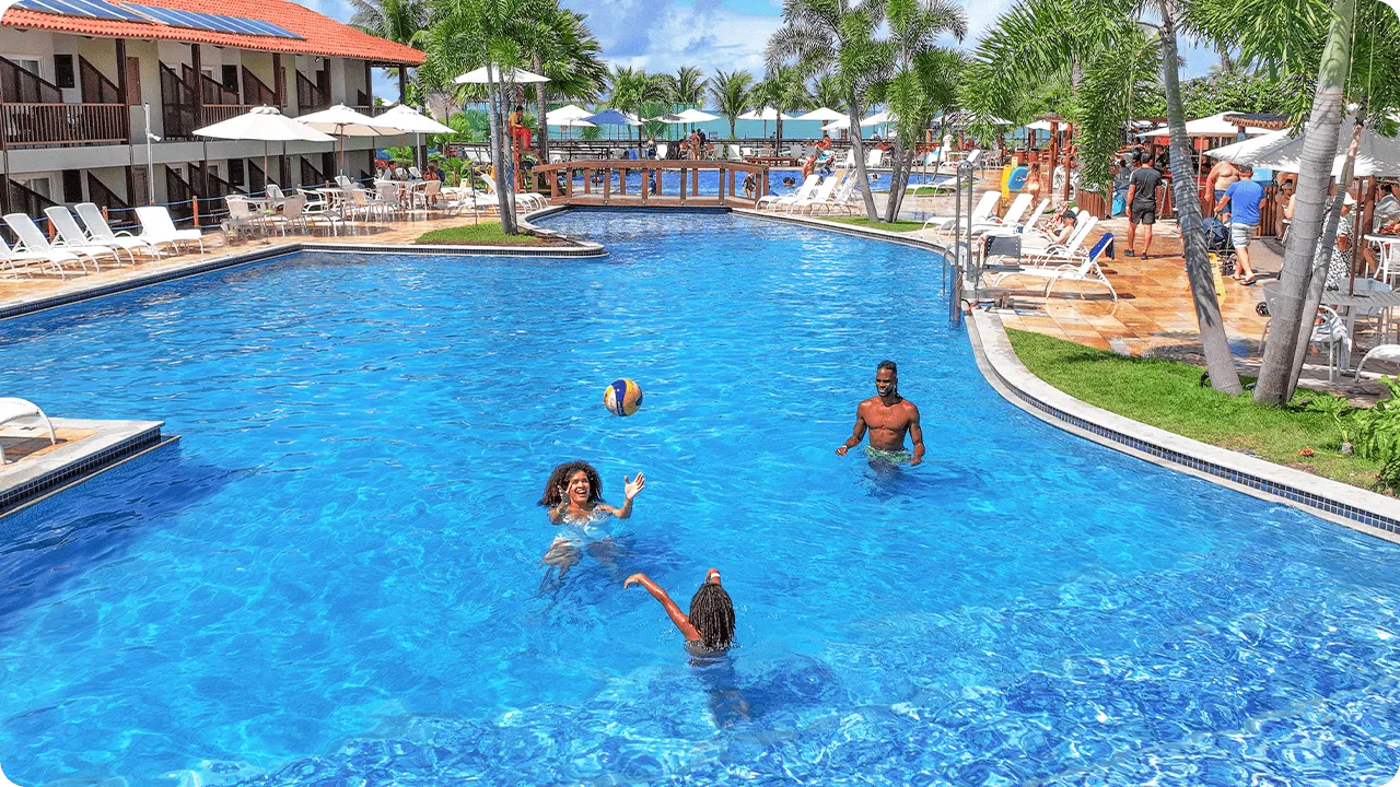 Una familia sonriente juega voleibol en la piscina azul de Salinas Maceió. La madre lanza la pelota al aire mientras padre e hija observan, en un día soleado rodeados de otros huéspedes y cocoteros.