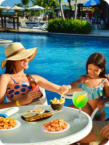 Una familia sonriente disfruta de bocadillos y bebidas en una mesa junto a la piscina en Salinas Maceió. Sobre la mesa hay porciones de camarones, papas fritas, un cóctel verde y una cerveza, un ejemplo de la experiencia todo incluido.
