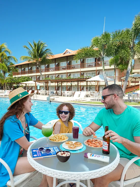 Una familia sonriente disfruta de bocadillos y bebidas en una mesa junto a la piscina en Salinas Maceió. Sobre la mesa hay porciones de camarones, papas fritas, un cóctel verde y una cerveza, un ejemplo de la experiencia todo incluido.