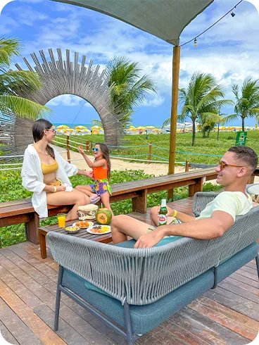 Família aproveitando o espaço externo do resort, sentada em sofás de madeira com vista para o mar e coqueiros, mesa com petiscos, coco e bebidas.