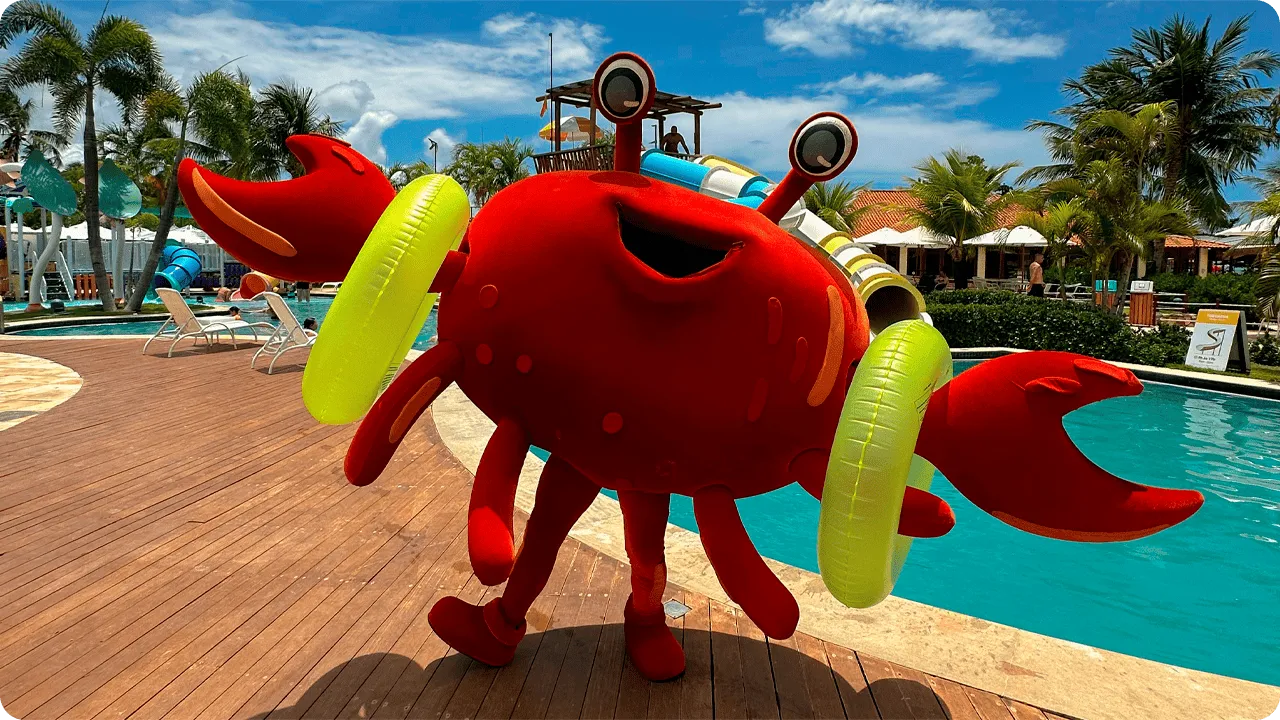 Mascote em formato de siri vermelho animado, com boias amarelas, ao lado da piscina do resort