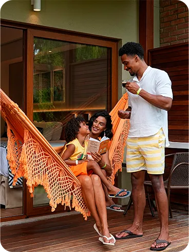 Família em momento de lazer na varanda privativa do quarto Standard Vitória-Régia. Mãe e filho leem um livro em uma rede laranja, enquanto o pai os observa sorrindo com uma xícara na mão.