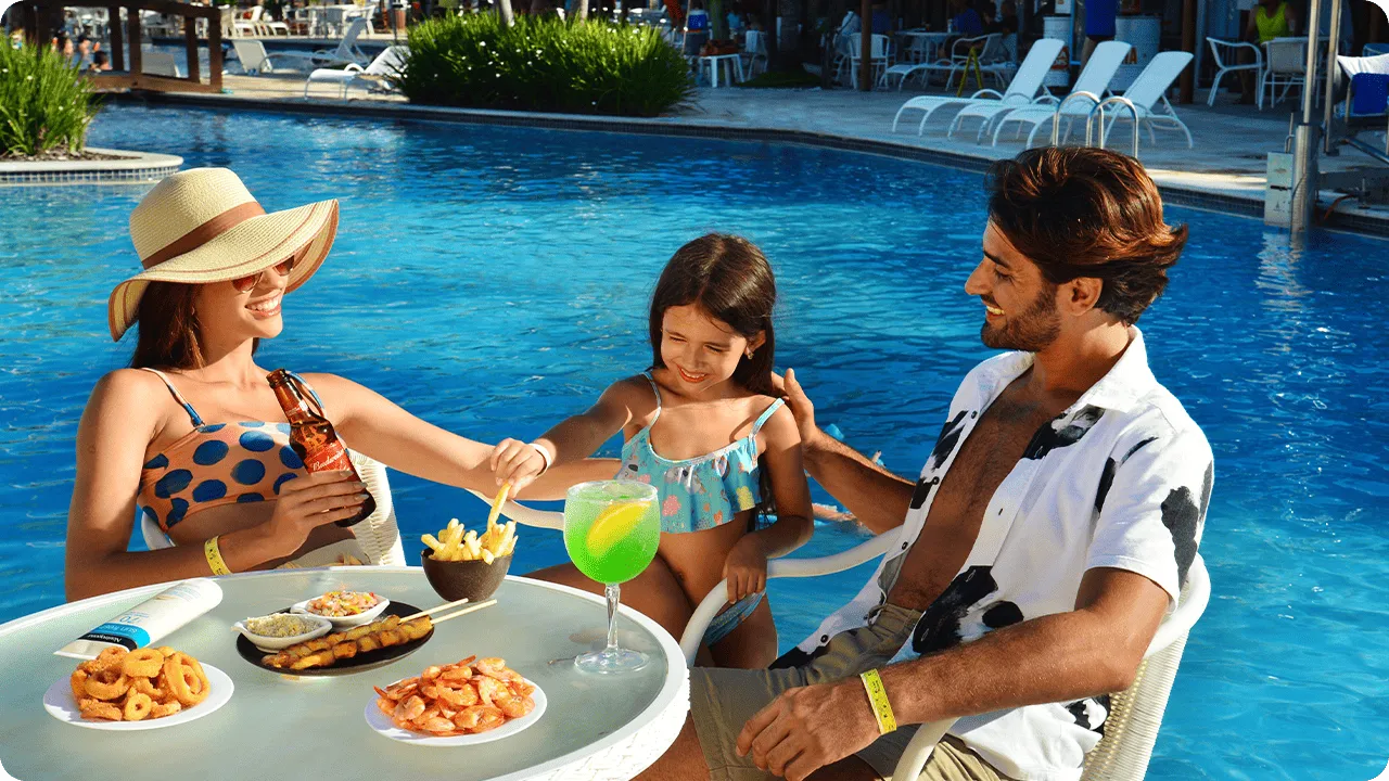 Una familia sonriente disfruta de bocadillos y bebidas en una mesa junto a la piscina en Salinas Maceió. Sobre la mesa hay porciones de camarones, papas fritas, un cóctel verde y una cerveza, un ejemplo de la experiencia todo incluido.