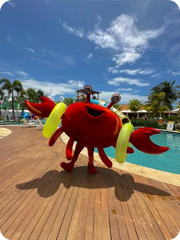 Mascote em formato de siri vermelho animado, com boias amarelas, ao lado da piscina do resort