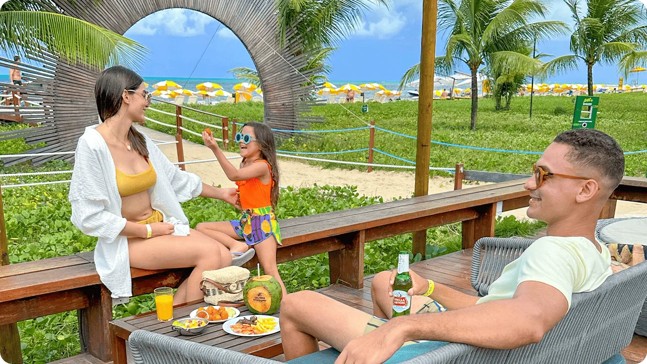Família aproveitando o espaço externo do resort, sentada em sofás de madeira com vista para o mar e coqueiros, mesa com petiscos, coco e bebidas.