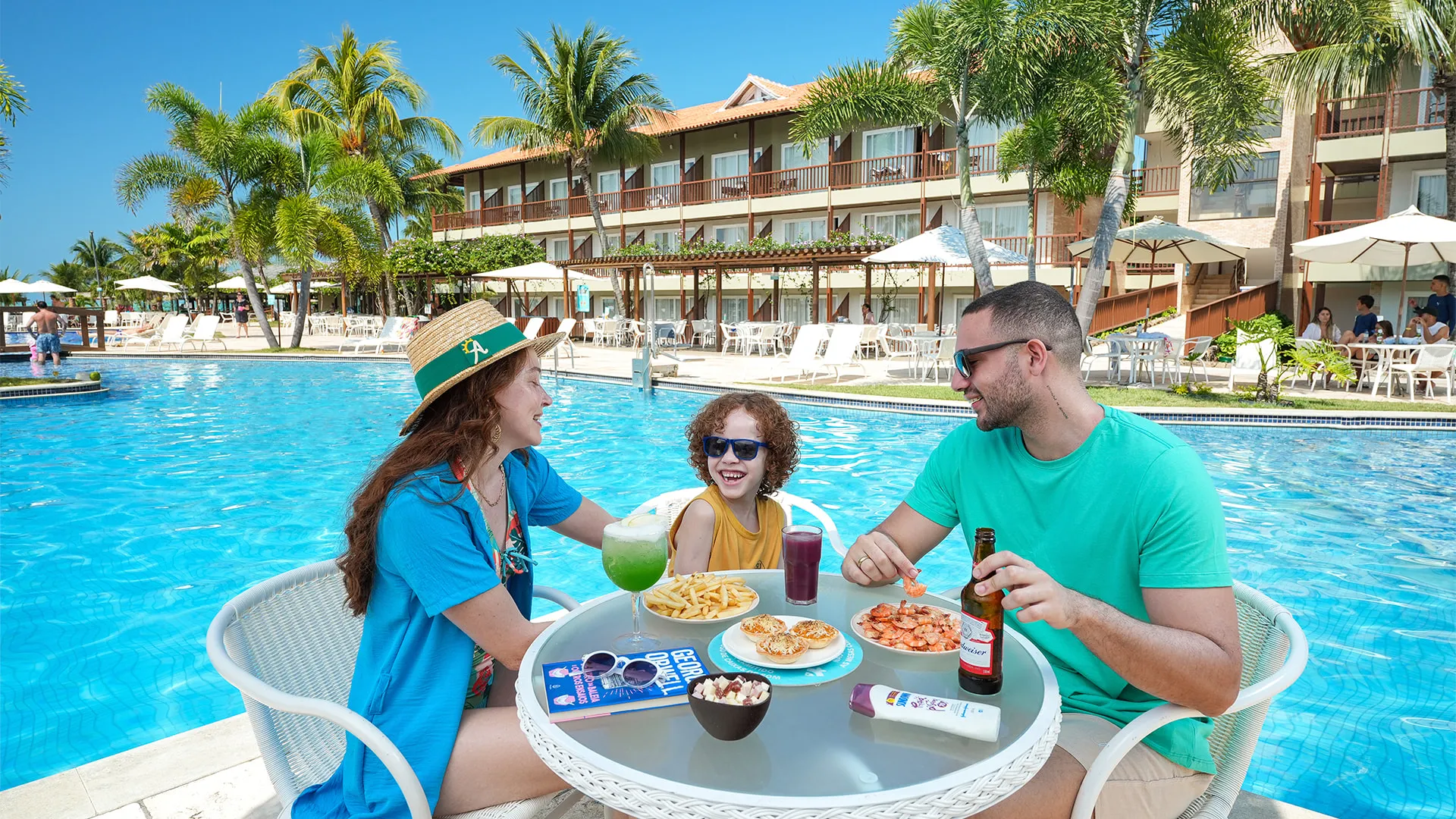 Una familia sonriente disfruta de bocadillos y bebidas en una mesa junto a la piscina en Salinas Maceió. Sobre la mesa hay porciones de camarones, papas fritas, un cóctel verde y una cerveza, un ejemplo de la experiencia todo incluido.
