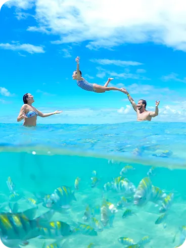 Foto dividida de las piscinas naturales de Maceió. En la superficie, una familia juega en el agua, mientras el padre lanza a su hija al aire. Bajo el agua, un gran banco de peces rayados nada en aguas cristalinas.