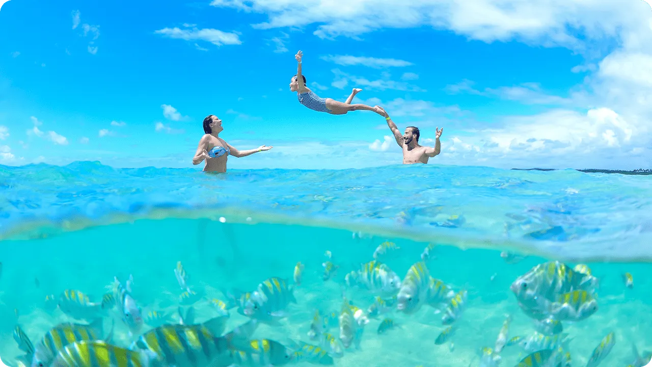 Foto dividida de las piscinas naturales de Maceió. En la superficie, una familia juega en el agua, mientras el padre lanza a su hija al aire. Bajo el agua, un gran banco de peces rayados nada en aguas cristalinas.