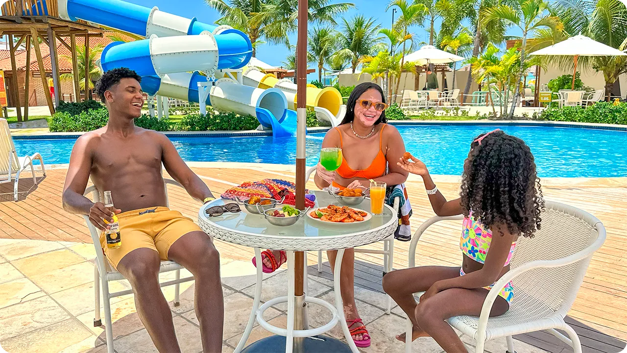 Padre, madre e hija disfrutan de un refrigerio en una mesa junto a la piscina del resort, con aperitivos y bebidas. Al fondo, el parque acuático con sus grandes toboganes azules y blancos completa la escena de vacaciones familiares.