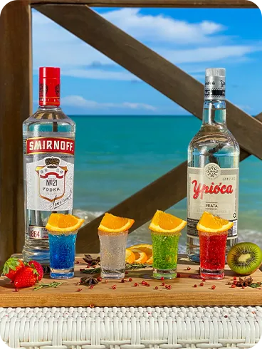 Garrafas de vodka e cachaça com copos de drinks coloridos decorados com laranja à beira da praia