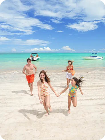 Família correndo sorridente na areia branca em frente ao mar azul cristalino com barcos ao fundo.
