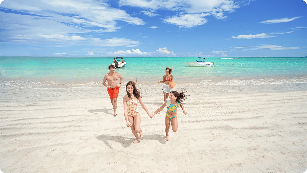Família correndo sorridente na areia branca em frente ao mar azul cristalino com barcos ao fundo.