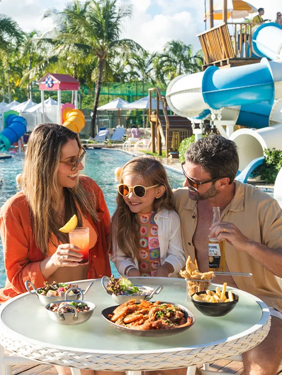 Padre, madre e hija disfrutan de un refrigerio en una mesa junto a la piscina del resort, con aperitivos y bebidas. Al fondo, el parque acuático con sus grandes toboganes azules y blancos completa la escena de vacaciones familiares.