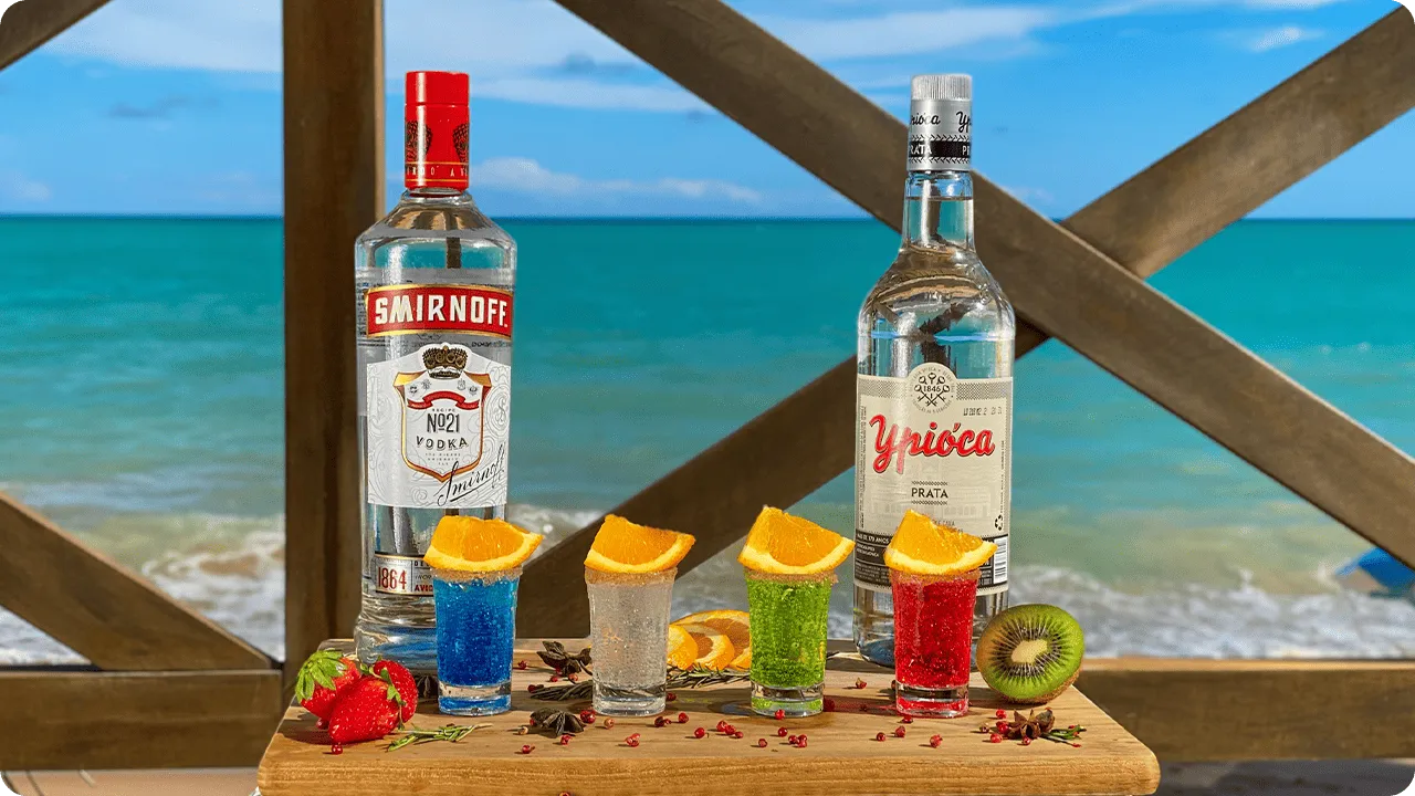 Garrafas de vodka e cachaça com copos de drinks coloridos decorados com laranja à beira da praia