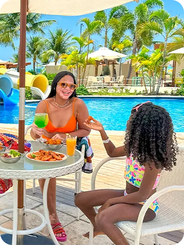 Padre, madre e hija disfrutan de un refrigerio en una mesa junto a la piscina del resort, con aperitivos y bebidas. Al fondo, el parque acuático con sus grandes toboganes azules y blancos completa la escena de vacaciones familiares.
