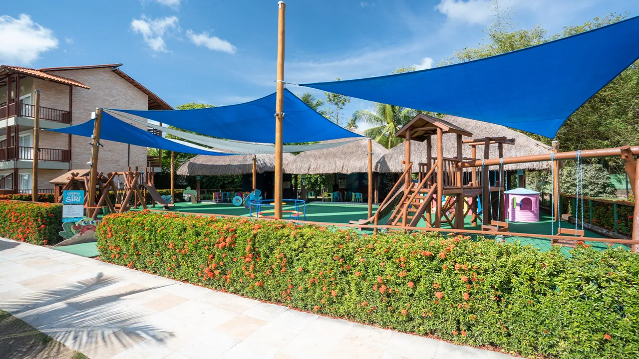 Área de lazer infantil, com diversos brinquedos pras crianças, alguns brinquedos tem uma cobertura azul pra proteger do sol. Na entrada tem um cercado de flores e ao fundo alguns bangalôs cobertos pras outras atividades.