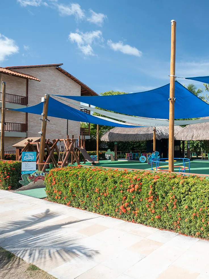 Área de lazer infantil, com diversos brinquedos pras crianças, alguns brinquedos tem uma cobertura azul pra proteger do sol. Na entrada tem um cercado de flores e ao fundo alguns bangalôs cobertos pras outras atividades.