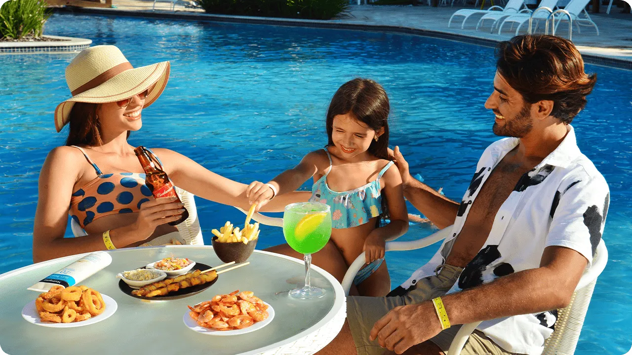 Família se diverte à beira da piscina com bebidas e petiscos variados em mesa de resort All Inclusive