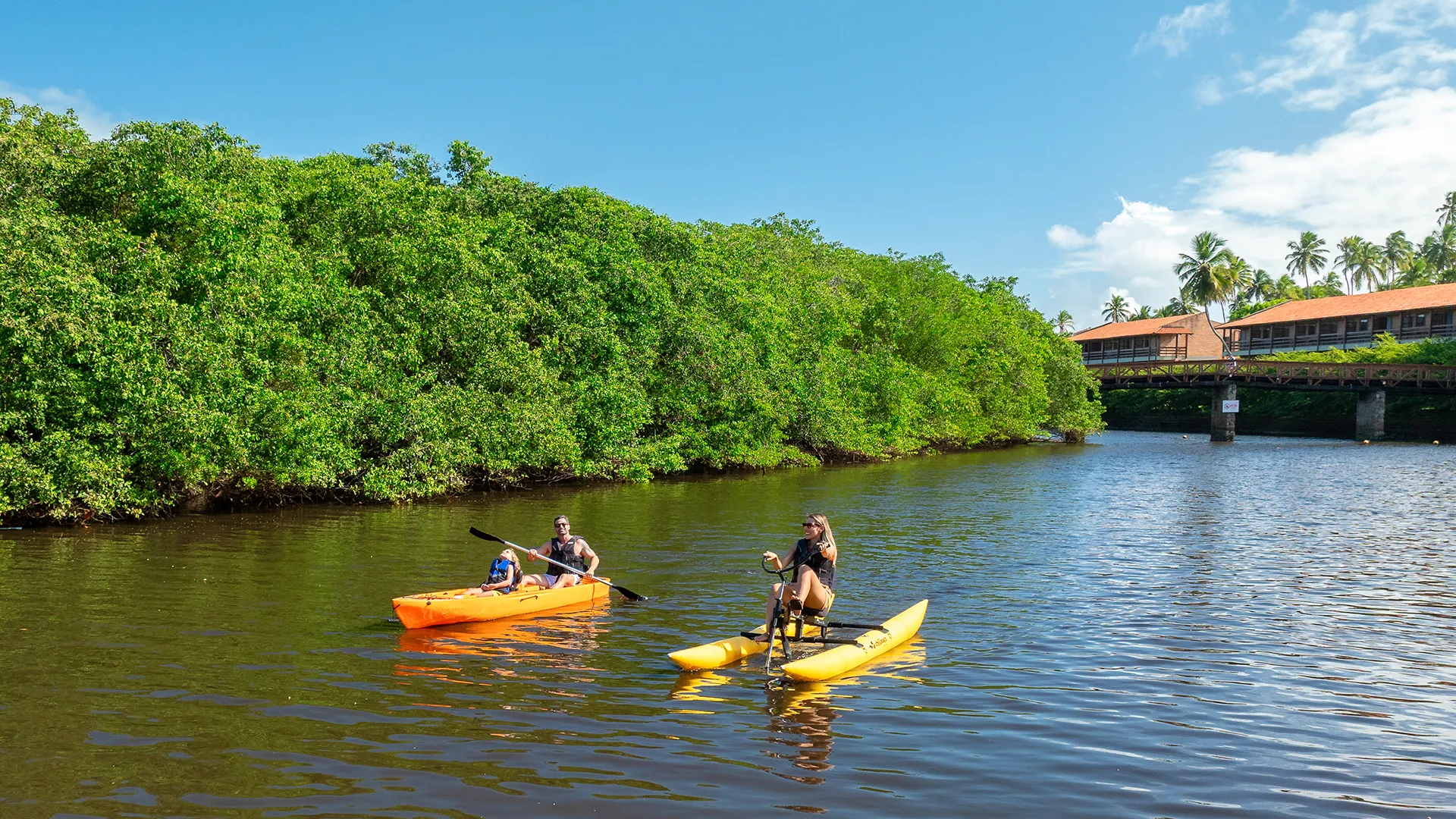Família praticando canoagem no rio que passa pelo meio do Salinas Maragogi, pai e filha estão num caiaque e mãe numa bicicleta aquática. Ao fundo plantas do manguezal, estrutura do resort e céu sem nuvens.
