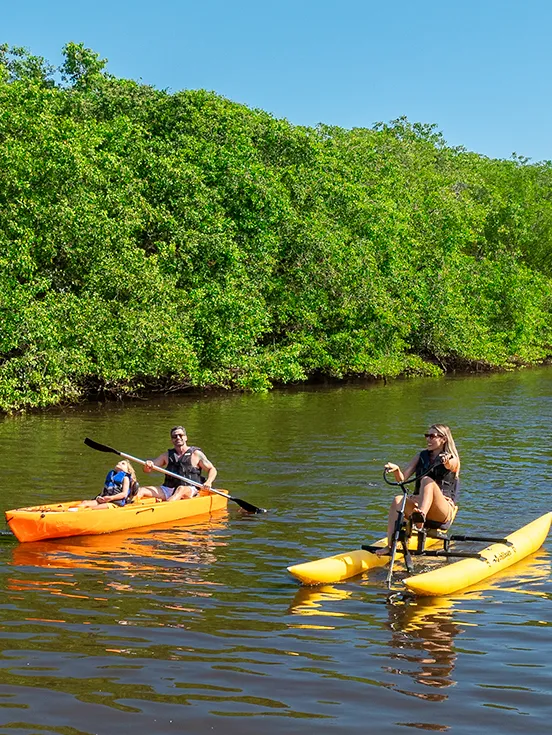 Família praticando canoagem no rio que passa pelo meio do Salinas Maragogi, pai e filha estão num caiaque e mãe numa bicicleta aquática. Ao fundo plantas do manguezal, estrutura do resort e céu sem nuvens.