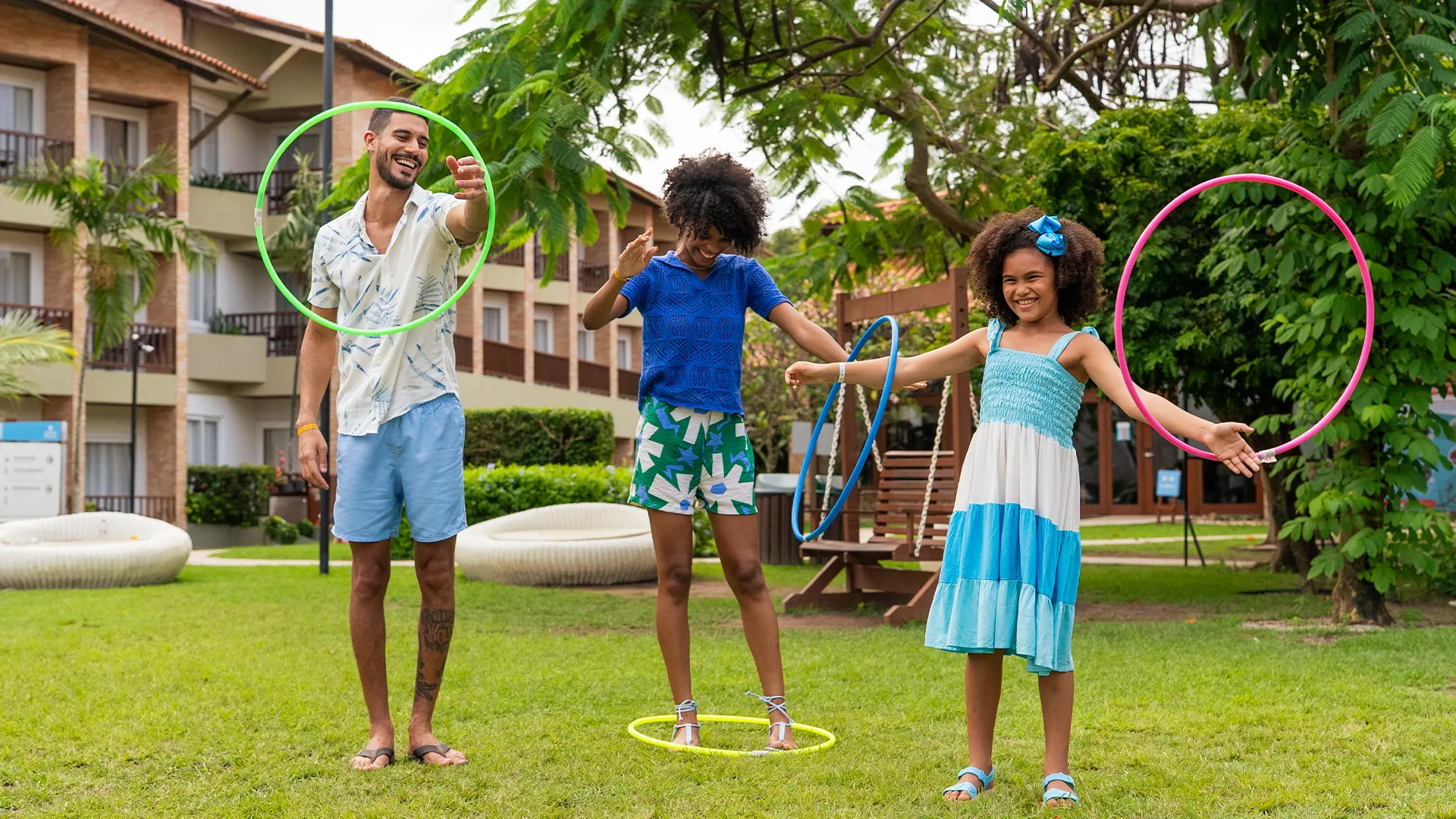 Família de pai, mãe e filha brincando com bambolê na área verde do Salinas Maceió, ao fundo os apartamentos do resort e muitas árvores.