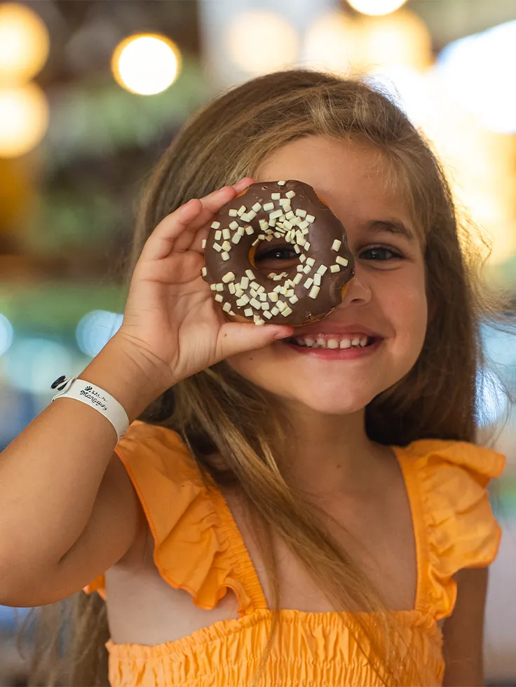 Criança sorridente com vestido laranja mostrando donuts de chocolate do resort Salinas Maragogi.