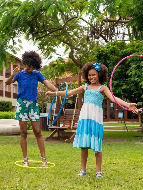 Família de pai, mãe e filha brincando com bambolê na área verde do Salinas Maceió, ao fundo os apartamentos do resort e muitas árvores.