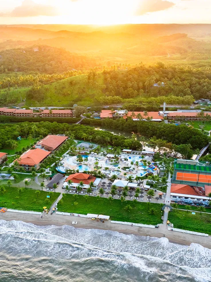 Imagem aérea do Salinas Maragogi mostrando o complexo de piscinas, dos quartos, pontos de consumo. Ao fundo e ao redor do resort grande imensidão de vegetação verde. Na parte debaixo da imagem pode se ve uma pequena faixa de areia e a parte da praia.