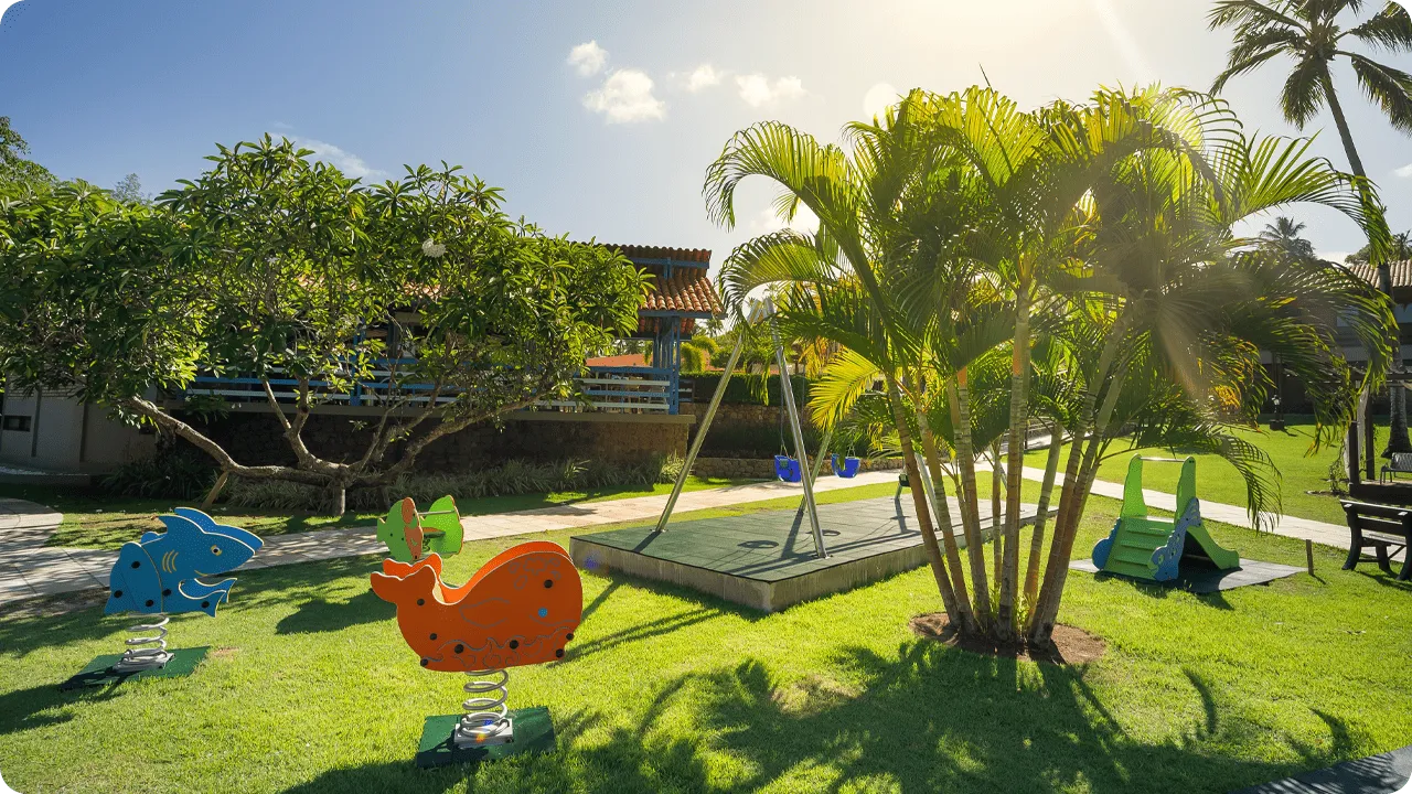 Área externa do resort com balanços e brinquedos coloridos para crianças em meio ao jardim.